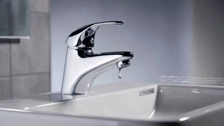 Beyaz seramik lavabo üzerinde krom renkli musluk ve ucunda asılı bir su damlası görülüyor.