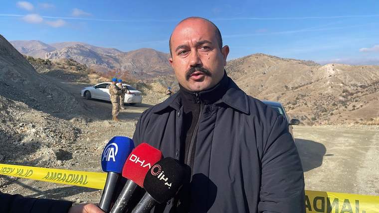 Kayalık dağlık arazide bir kişi, çeşitli televizyon mikrofonlarına konuşurken arkasında güvenlik şeridi görülüyor.