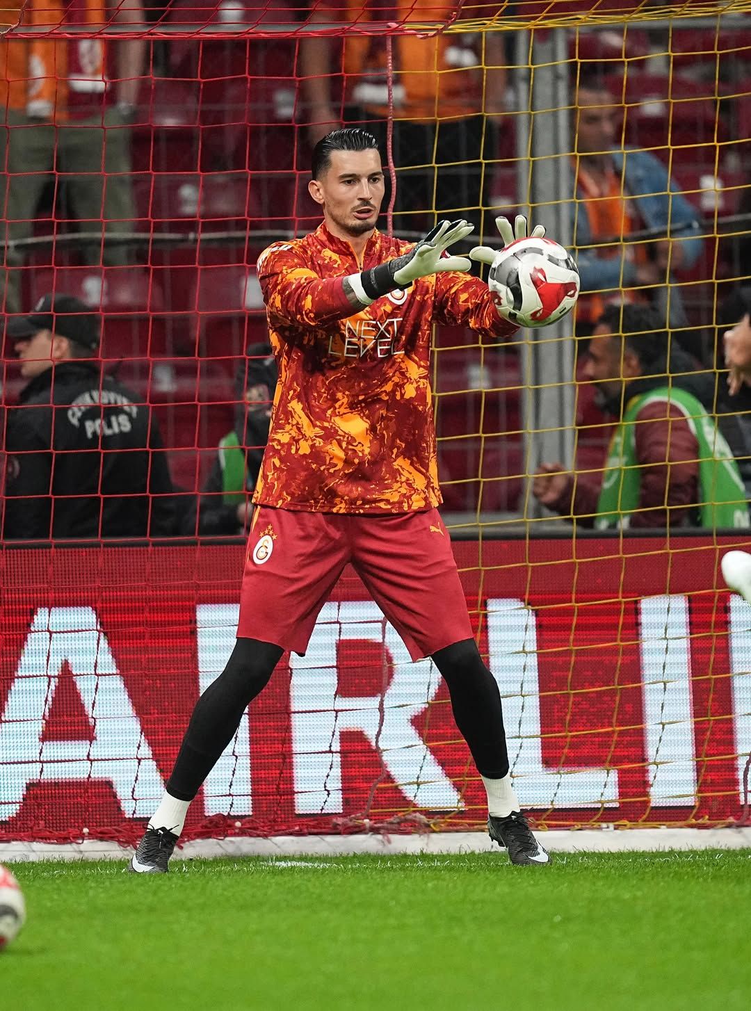 Galatasaray armalı forması ve eldivenleri görülen kaleci, kale önünde topu elleriyle tutuyor.
