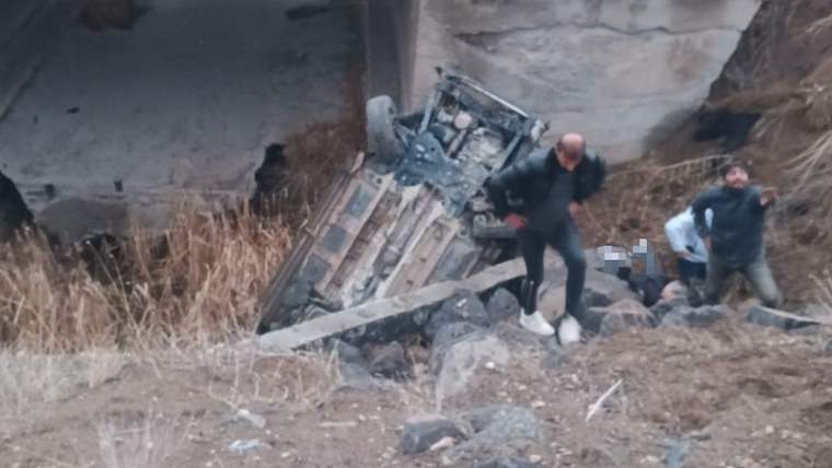 Beton köprü ayağının yanında ters dönmüş araç enkazı ve enkazın çevresinde duran iki kişi görülüyor.