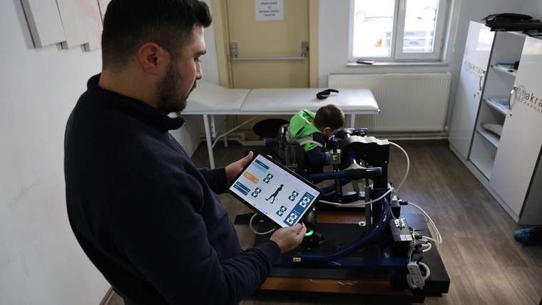 Bir kişi, mobil yürüme robotunu kontrol eden bir tablet tutuyor; arka planda oturan bir çocuk görünüyor.