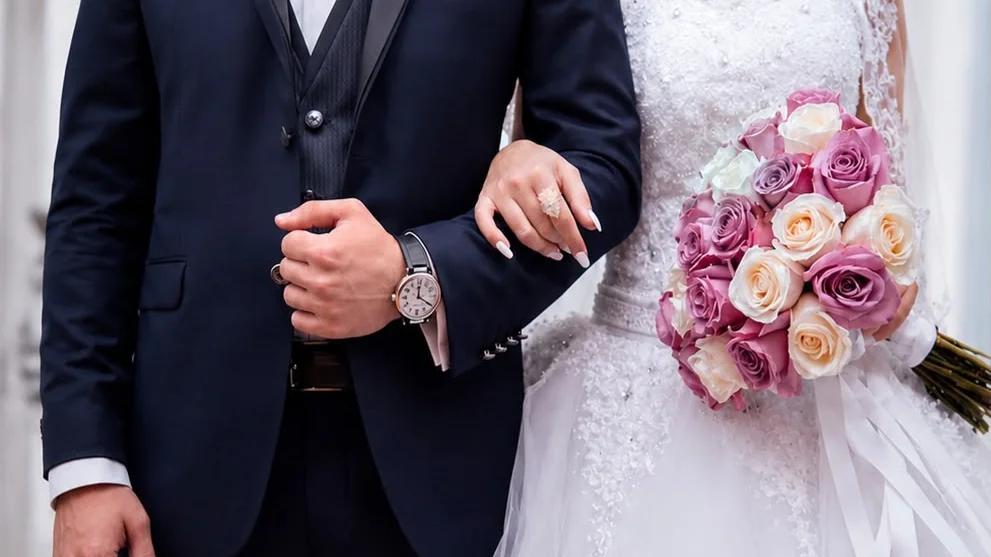 Damat, siyah takım elbise giymiş, gelin ise beyaz gelinlik içinde, elinde çiçek buketi tutuyor.