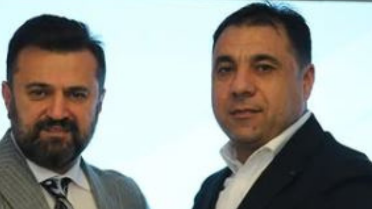 İki kişi, bir arada poz veriyor; biri takım elbise giymekte, diğeri ise ceketli.