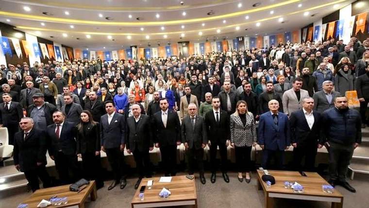 Konferans salonunda, ön sırada yan yana duran kişiler ve arkalarında kalabalık bir topluluk görülüyor.