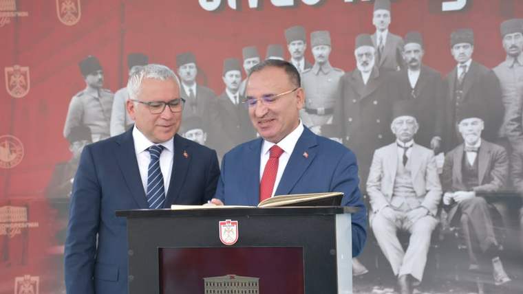 Kürsü önünde duran iki erkek, açık bir dosyaya bakıyor; arkalarında tarihi kişilerin portreleri bulunuyor.