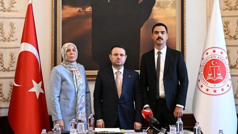 Türk bayrağı ve Adalet Bakanlığı logolu bayrak önünde üç kişi, masadaki mikrofonla birlikte duruyor.