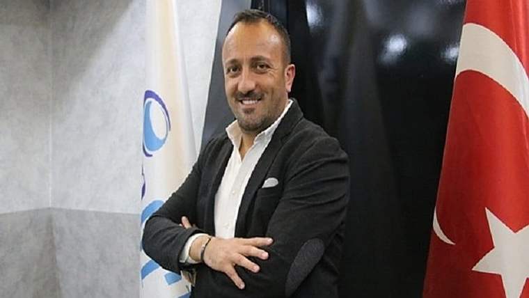 Kollarını kavuşturmuş bir adam, gülümseyerek poz veriyor. Arkada Türk bayrağı ve başka bir bayrak görünmekte.
