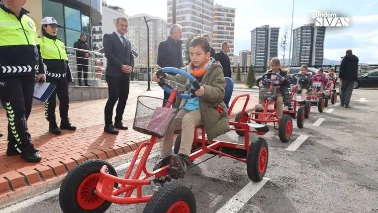Önde bir çocuk kırmızı pedallı araca binerken, arkasında başka çocuklar ve reflektörlü iki görevli görülüyor.