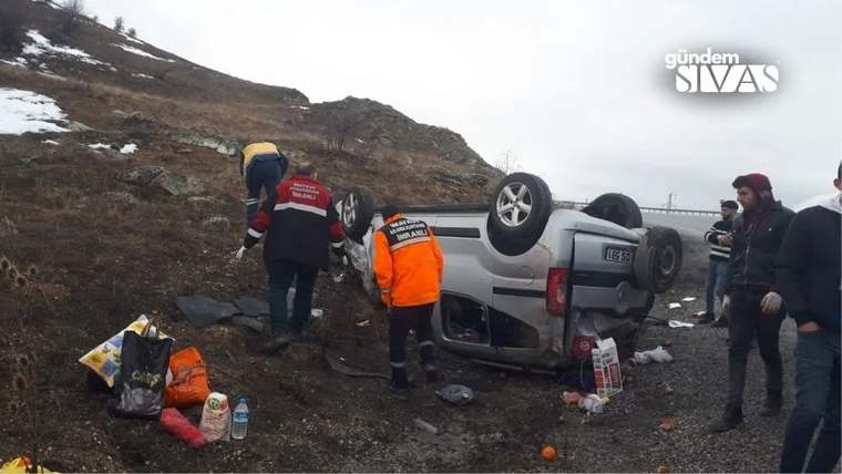 Kayalık yol kenarında ters dönmüş otomobilin çevresinde turuncu yelekli kişiler ve birkaç kişi duruyor.