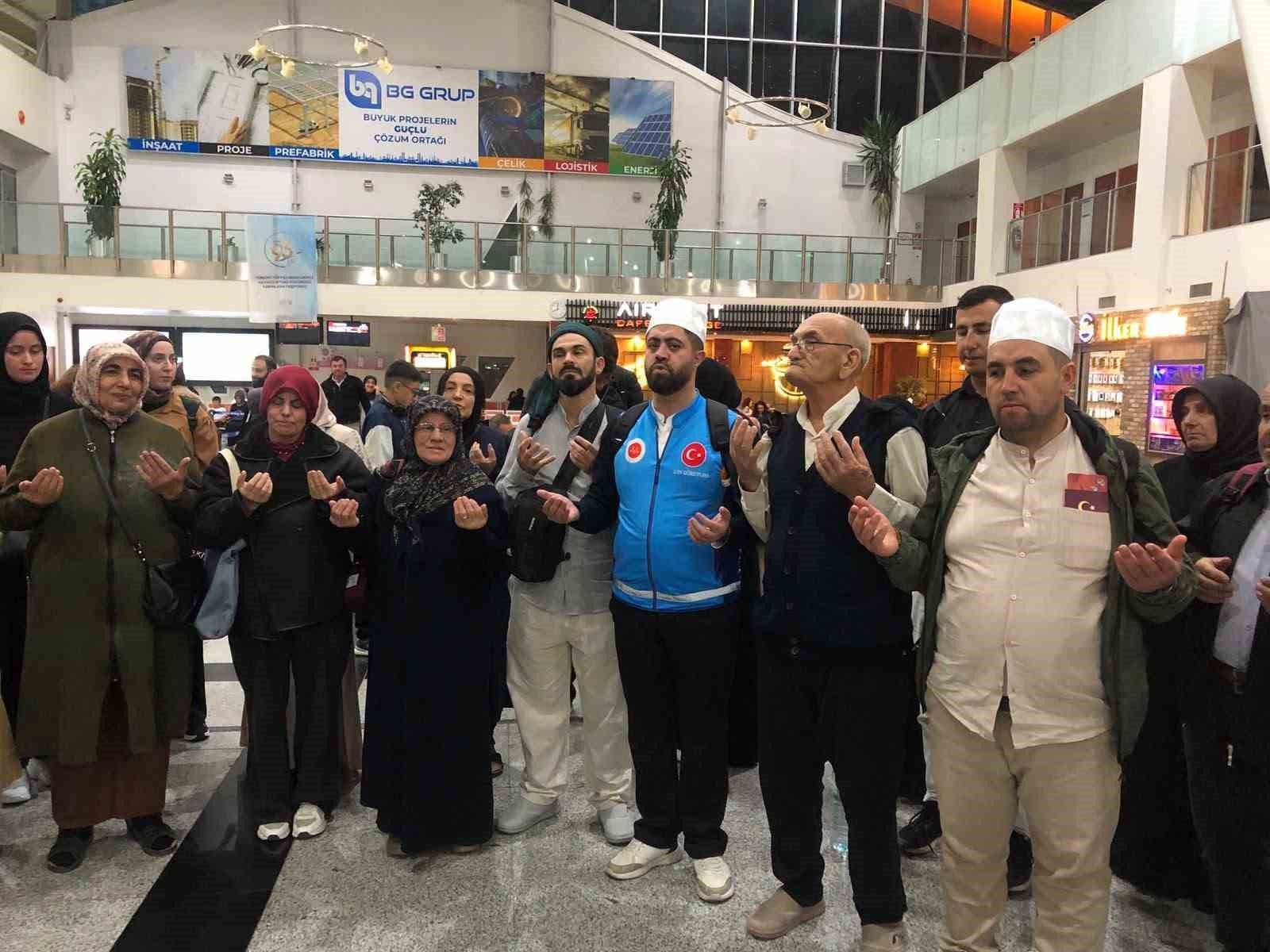 Terminal salonunda, ellerini dua eder gibi kaldırmış kadın ve erkeklerden oluşan bir grup duruyor.