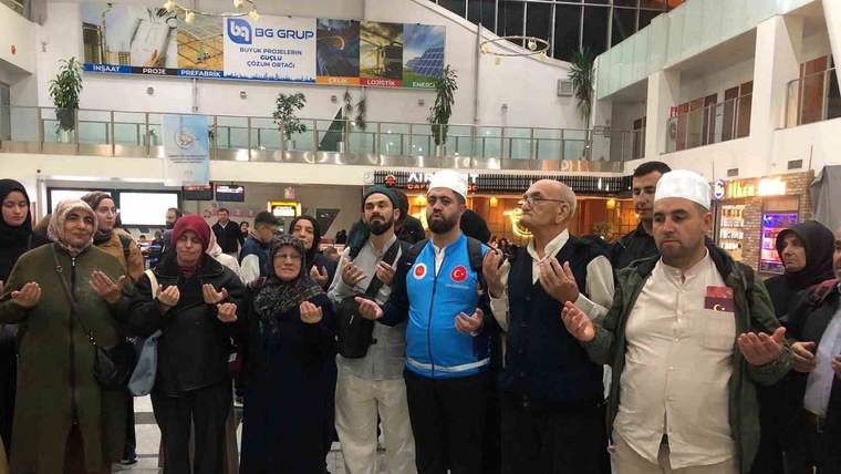 Terminal salonunda, ellerini dua eder gibi kaldırmış kadın ve erkeklerden oluşan bir grup duruyor.