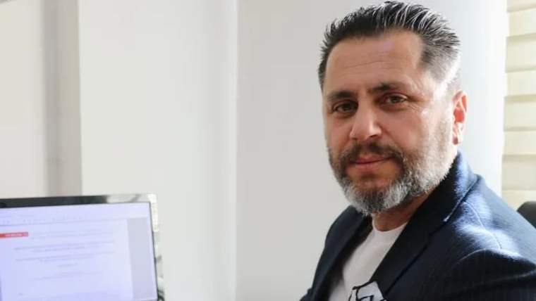 Bir kişi, bilgisayar ekranının önünde oturuyor, gülümseyerek kameraya bakıyor.