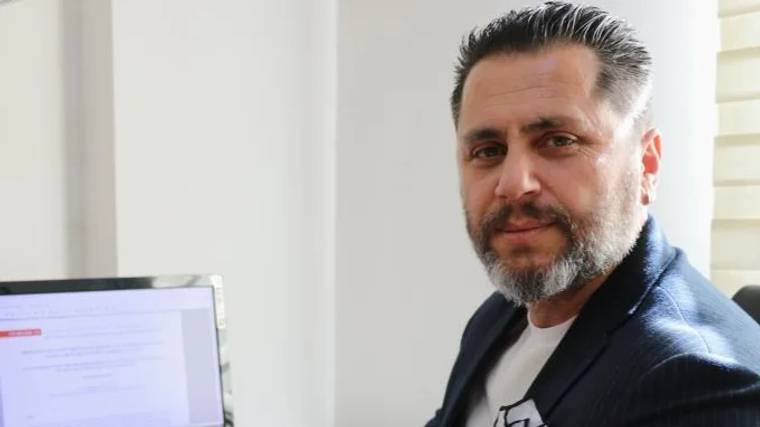 Bir kişi, bilgisayar ekranının önünde oturuyor, gülümseyerek kameraya bakıyor.