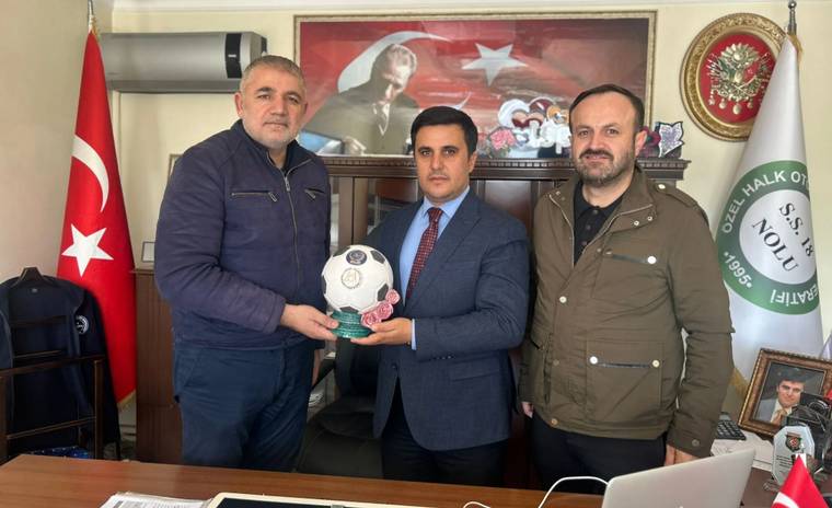 Bir ofiste üç erkek, Türk bayrağı önünde futbol topunu birlikte tutuyor; masada bilgisayar bulunuyor.