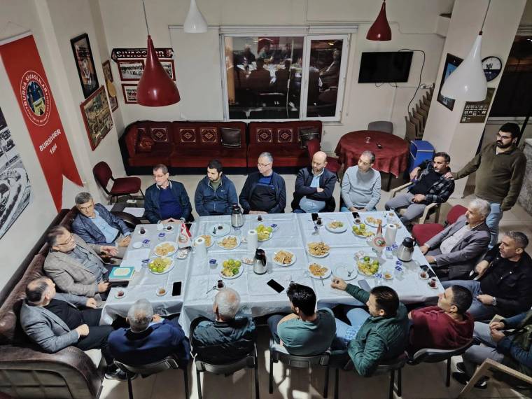 Bir odada uzun masa çevresinde oturan kalabalık grup, masadaki yiyecekler ve duvarlardaki süslemelerle görülüyor.