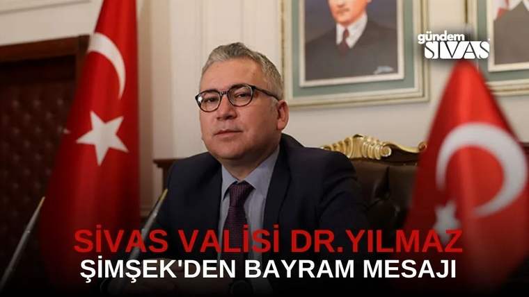 Masa başında oturan gözlüklü bir kişi, iki Türk bayrağı ve duvarındaki portrelerin önünde görülüyor.