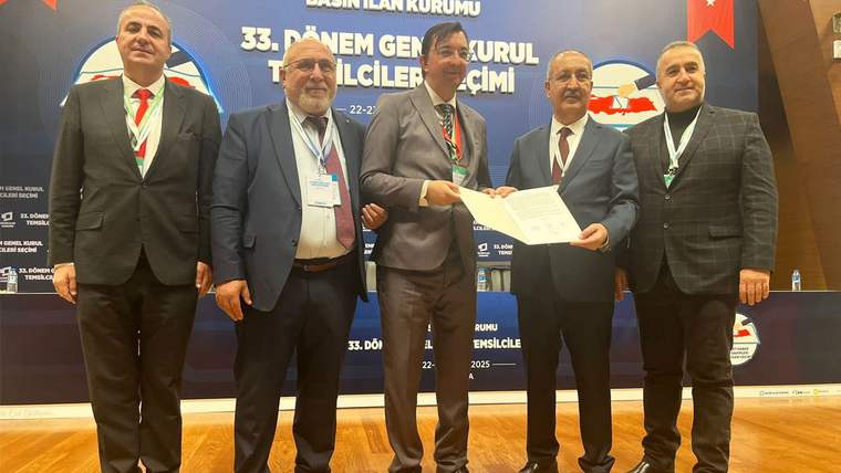 Kürsü önünde beş erkek yan yana duruyor; ortadaki iki kişi bir belgeyi birlikte tutuyor.