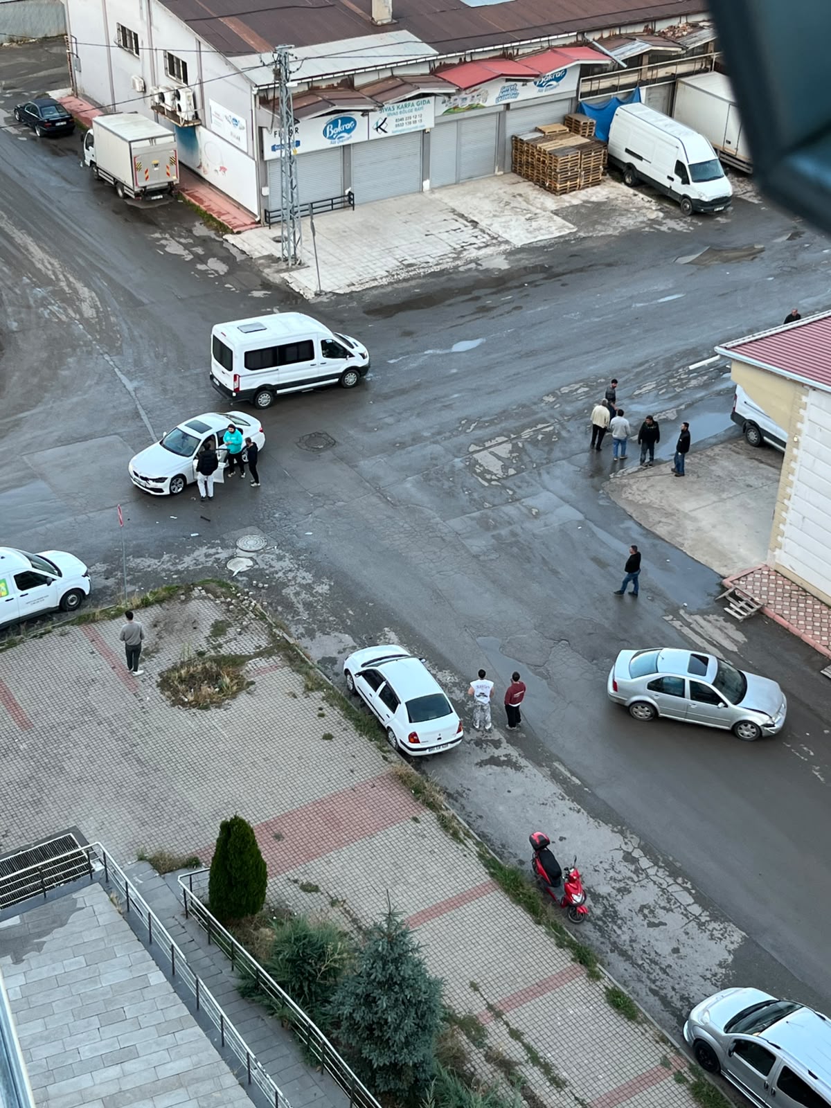 Kavşakta çeşitli araçlar ve yayalar görülürken, beyaz bir otomobilin yanında birkaç kişi bekliyor.