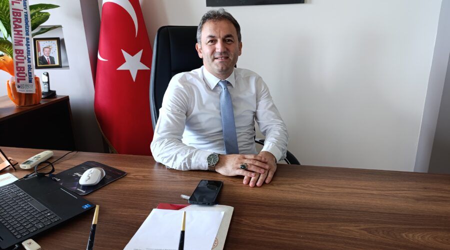 Masasında oturan takım elbiseli bir kişi, arkasındaki Türk bayrağı ve çalışma masasındaki eşyalarla görülüyor.