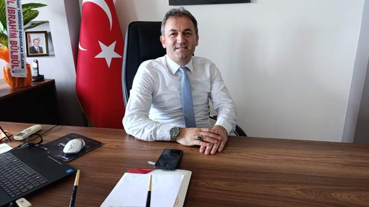 Masasında oturan takım elbiseli bir kişi, arkasındaki Türk bayrağı ve çalışma masasındaki eşyalarla görülüyor.