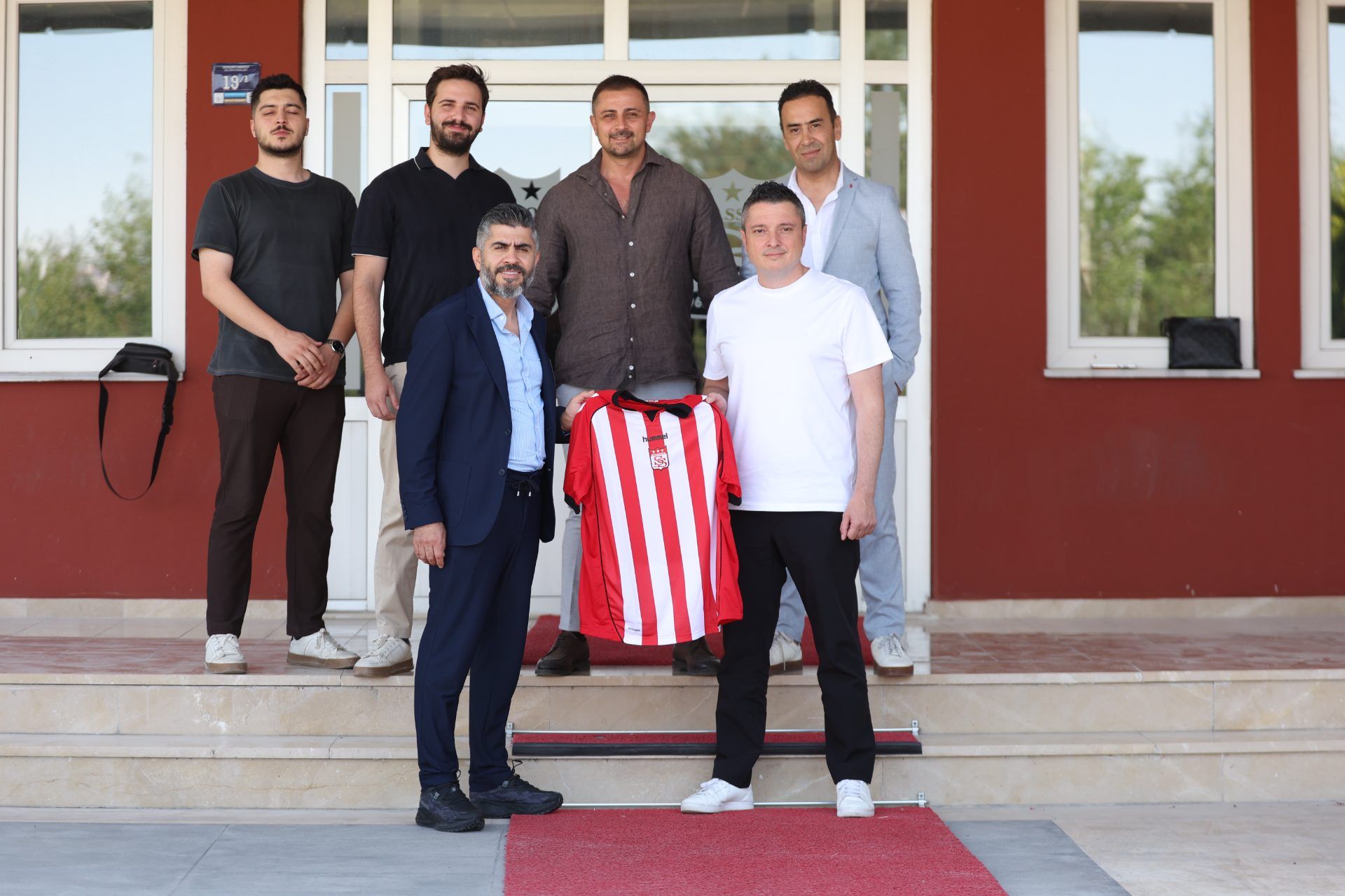 Bir bina girişinde altı kişi, önlerinde kırmızı-beyaz çizgili bir forma ile birlikte duruyor.