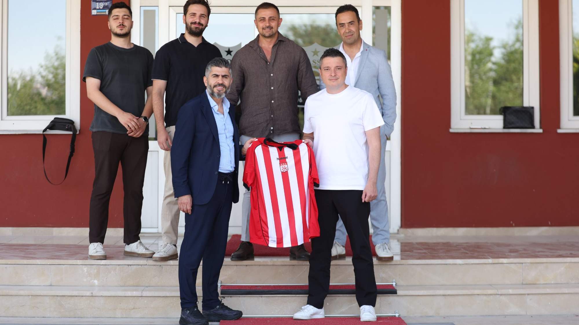 Bir bina girişinde altı kişi, önlerinde kırmızı-beyaz çizgili bir forma ile birlikte duruyor.