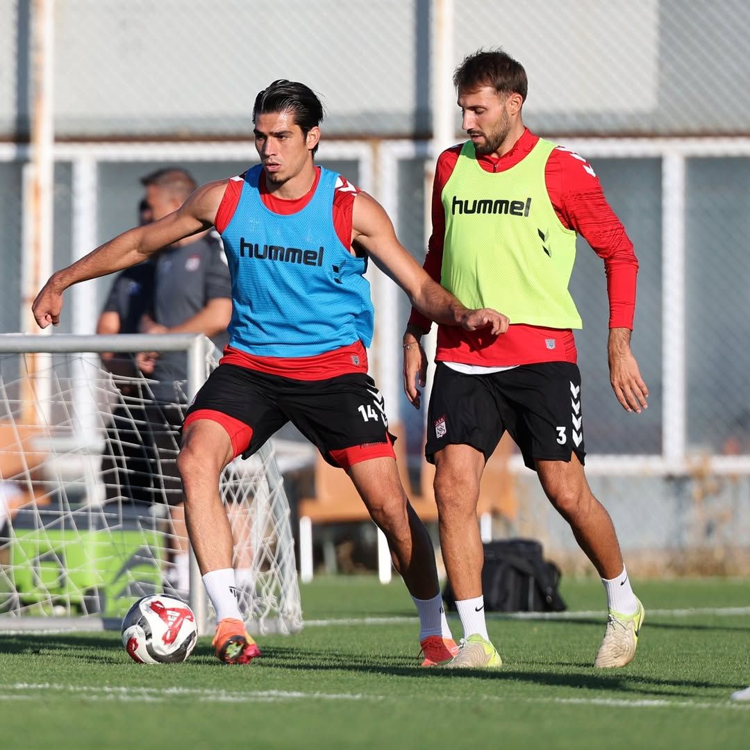 İki futbolcu, antrenman sahasında top için mücadele ederken biri ayağıyla topa dokunuyor.