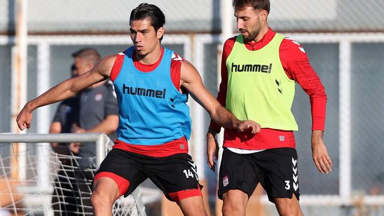 İki futbolcu, antrenman sahasında top için mücadele ederken biri ayağıyla topa dokunuyor.