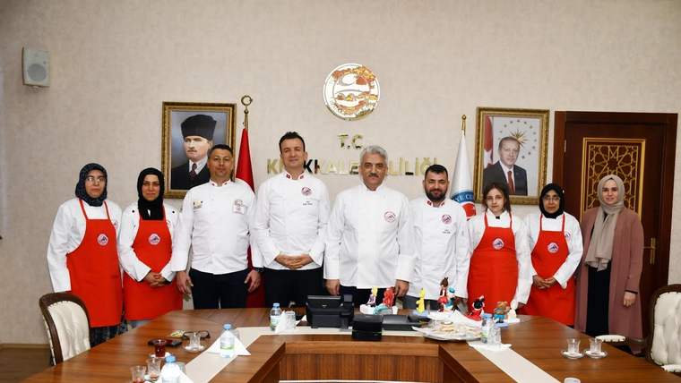 Masa çevresinde beyaz aşçı kıyafetleri ve kırmızı önlükler giyen bir grup kişi birlikte duruyor.