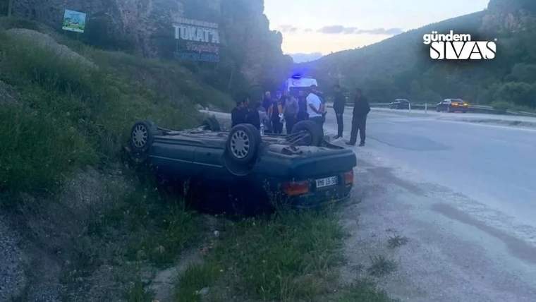 Kırsal yolda ters dönen bir otomobilin çevresinde birkaç kişi duruyor; arkada mavi ışık görülüyor.
