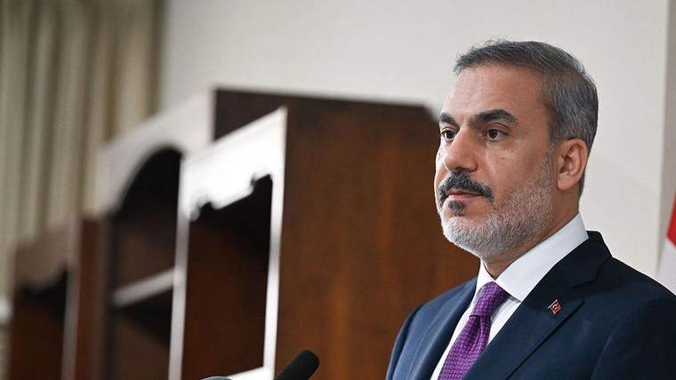 Kravatlı bir kişi, ahşap kürsünün önünde mikrofonla konuşurken başını yana çevirmiş görünüyor.