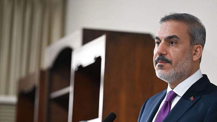 Kürsüde konuşan bir kişi, arka planda ahşap bir raf görünmekte.