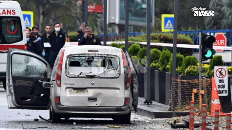 Arka camı kırılmış beyaz otomobilin kapısı açık; çevresinde polis şeridi, dubalar ve yayalar görülüyor.