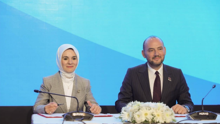Mavi ekran önünde, masa başında oturan başörtülü bir kadın ve erkek konuşmacının önünde mikrofonlar ile çiçekler bulunuyor.