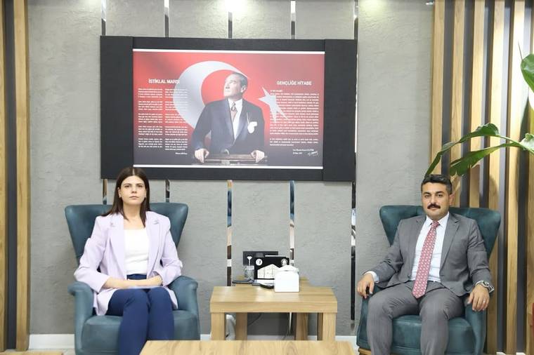 İç mekânda iki kişi koltuklarda oturuyor; arkalarında Atatürk portresi ve kırmızı-siyah bir pano bulunuyor.