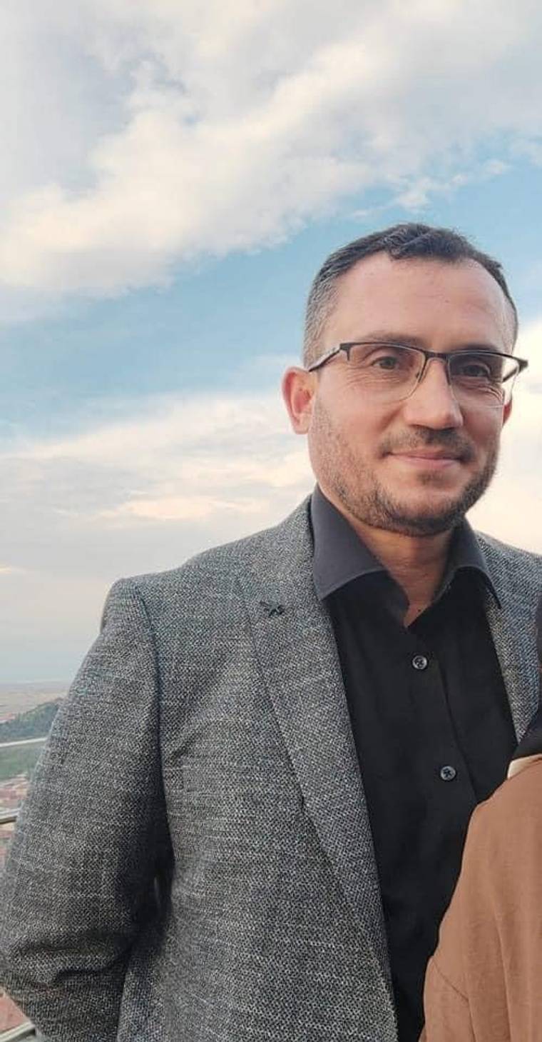 Gözlüklü bir adam, gri ceket ve siyah gömlekle açık havada kameraya dönük duruyor.