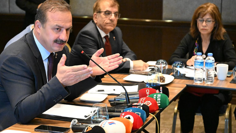 Bir kişi, masanın arkasında el hareketleriyle konuşuyor; masada su şişeleri ve mikrofonlar var.
