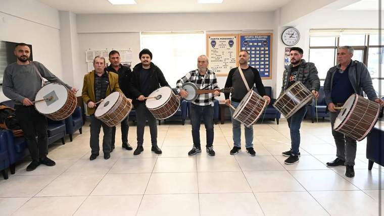 Bir grup erkek, iç mekânda ellerindeki farklı boyutlardaki davullarla yan yana duruyor.