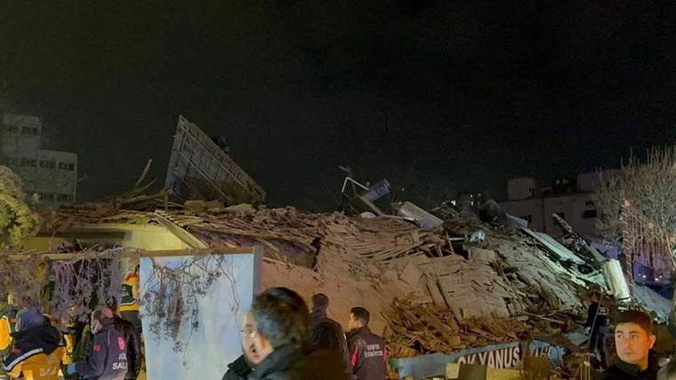 Gece çöken binanın enkazı önünde, baretli ve reflektif kıyafetli kişiler çalışma yapıyor.