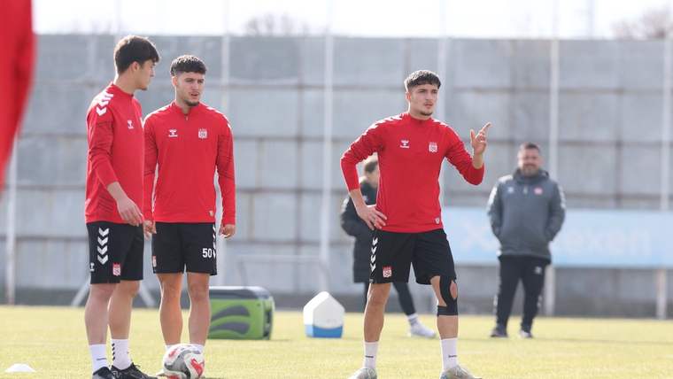 Kırmızı forma giymiş üç futbolcu antrenman yapıyor, biri elini işaret ediyor.