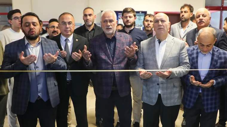 Bir grup insan, kapalı alanda sarı kurdele önünde dururken ortadaki kişi ellerini kaldırıyor.