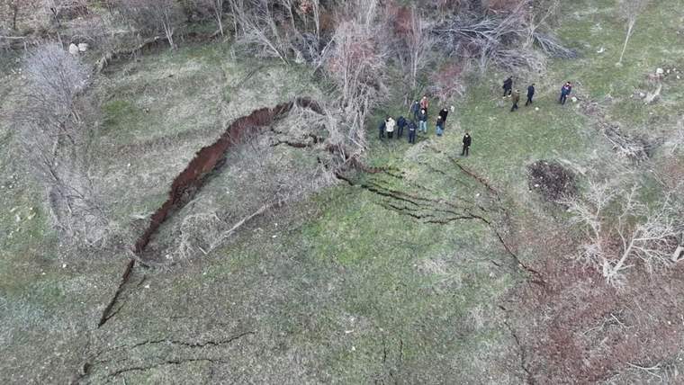 Ağaçlık ve çimenlik alanda uzanan derin yarığın çevresinde bir grup kişi ayakta duruyor.
