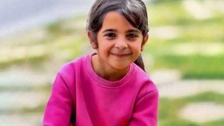 Açık havada, pembe kazak giyen küçük bir kız çocuğu kameraya bakarak gülümsüyor.