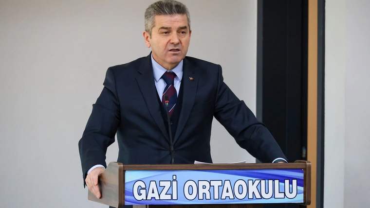 Kravatlı bir kişi, üzerinde “GAZİ ORTAOKULU” yazan kürsüde konuşma yaparken görülüyor.