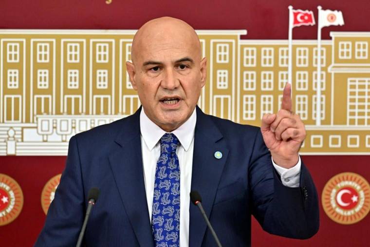 Kürsüde konuşan kel bir erkek, sağ işaret parmağını kaldırmış; arkasında bina çizimi ve Türk bayrakları var.