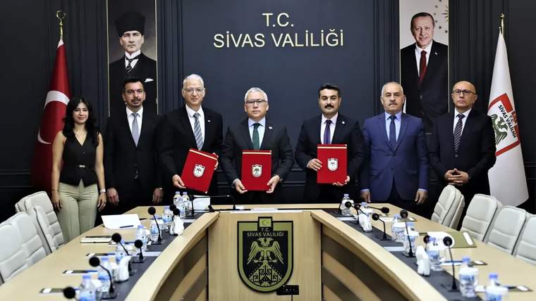 Sivas Valiliği yazılı salonda, toplantı masası çevresinde duran yedi kişi ve ellerindeki kırmızı dosyalar görülüyor.