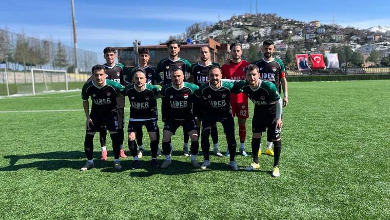 Futbol sahasında siyah formalı on bir oyuncu takım fotoğrafı için poz veriyor; arkada kale ve evler görülüyor.