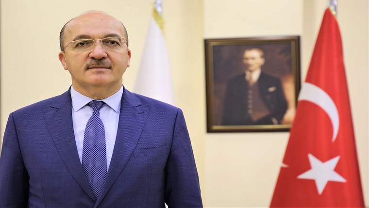 Takım elbiseli bir kişi, duvarda çerçeveli portre ve Türk bayrağının bulunduğu odada duruyor.