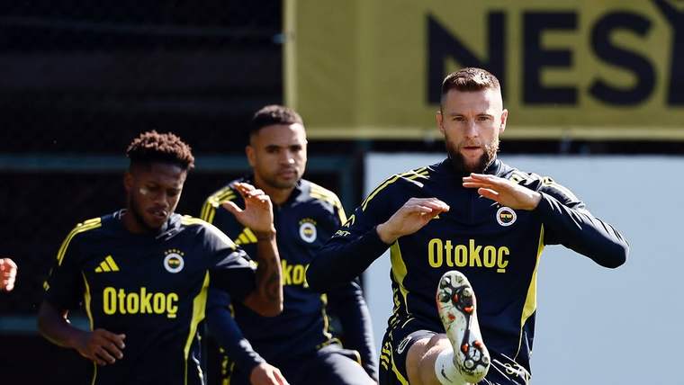 Antrenman sahasında üç futbolcu koşu çalışması yaparken öndeki oyuncu dizini kaldırıyor; çevrede renkli koniler bulunuyor.