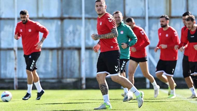 Çim sahada kırmızı formalar giyen futbolcular koşu antrenmanı yaparken, bir futbolcu top sürüyor.